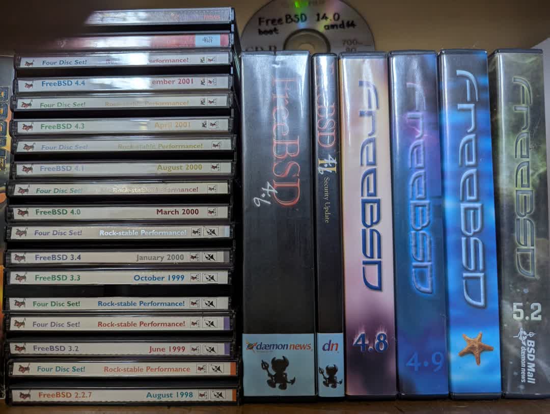 Photograph of a collection of CD and DVD cases for FreeBSD releases from 2.2.7 in August 1998 to 5.2. Behind these is a FreeBSD 14.0 amd64 labeled CD-R.